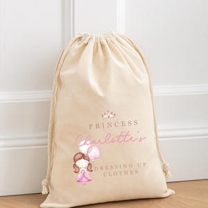 May include: A beige drawstring bag with the text "Princess Charlotte's Dressing Up Clothes" in pink and brown. The bag features a watercolor illustration of a princess holding balloons and a crown.