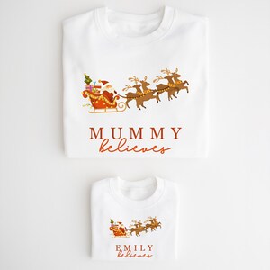 May include: Two white t-shirts with a Christmas theme. The larger shirt says "MUMMY believes" and the smaller shirt says "EMILY believes". Both shirts feature a cartoon image of Santa Claus in his sleigh pulled by reindeer.