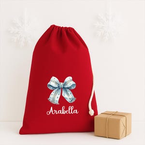 Puede incluir: Una bolsa de regalo roja con cord&oacute;n, con un dise&ntilde;o de lazo en acuarela en tonos azules y grises, y el nombre "Arabella" en escritura cursiva. Un peque&ntilde;o paquete marr&oacute;n envuelto est&aacute; al lado de la bolsa. La bolsa tiene un cierre de cord&oacute;n blanco.