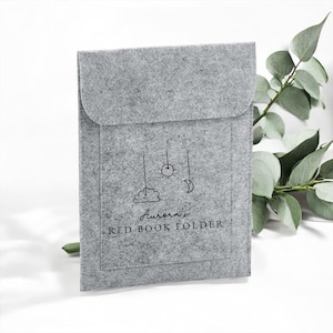May include: A grey felt book folder with a flap closure. The front has a whimsical design of a cloud, sun, and moon hanging from strings, with the text "Aurora's RED BOOK FOLDER" below. The folder is rectangular and has a pocket.