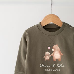 May include: A green sweatshirt with a brown bear and a smaller brown bear holding hands. The text "Mama & Ellie since 2022" is printed on the sweatshirt.