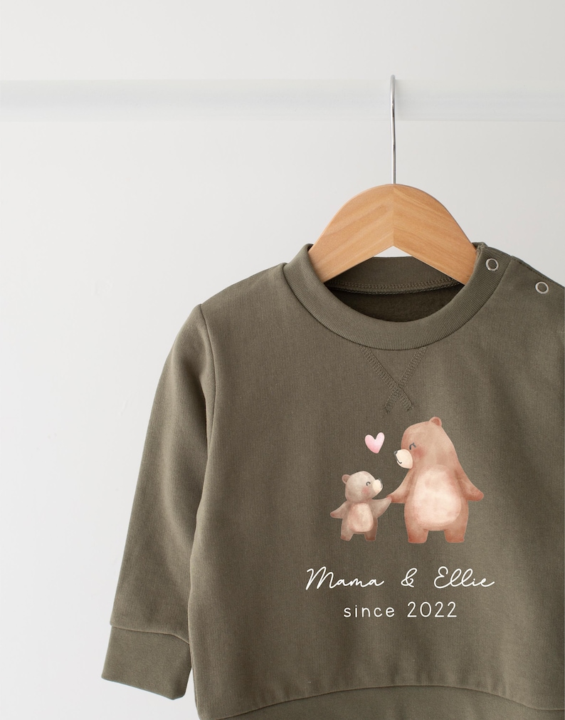 May include: A green sweatshirt with a brown bear and a smaller brown bear holding hands. The text "Mama & Ellie since 2022" is printed on the sweatshirt.