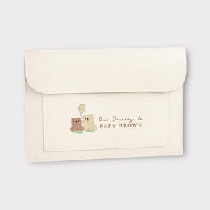 May include: Cream-coloured envelope-style pouch with a flap closure. A smaller pocket on the front features an illustration of two teddy bears and a balloon. The text "Our Journey to BABY BROWN" is printed in brown.