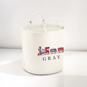May include: White canvas storage bin with a red, blue and grey train design and the name "GRAY" printed on the front.