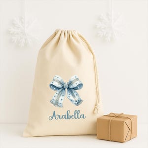 Puede incluir: Una bolsa de regalo beige con cord&oacute;n, con un dise&ntilde;o de lazo azul y el nombre "Arabella" impreso en azul. Una peque&ntilde;a caja de regalo envuelta est&aacute; a la derecha. Dos decoraciones de copos de nieve blancos cuelgan en el fondo.