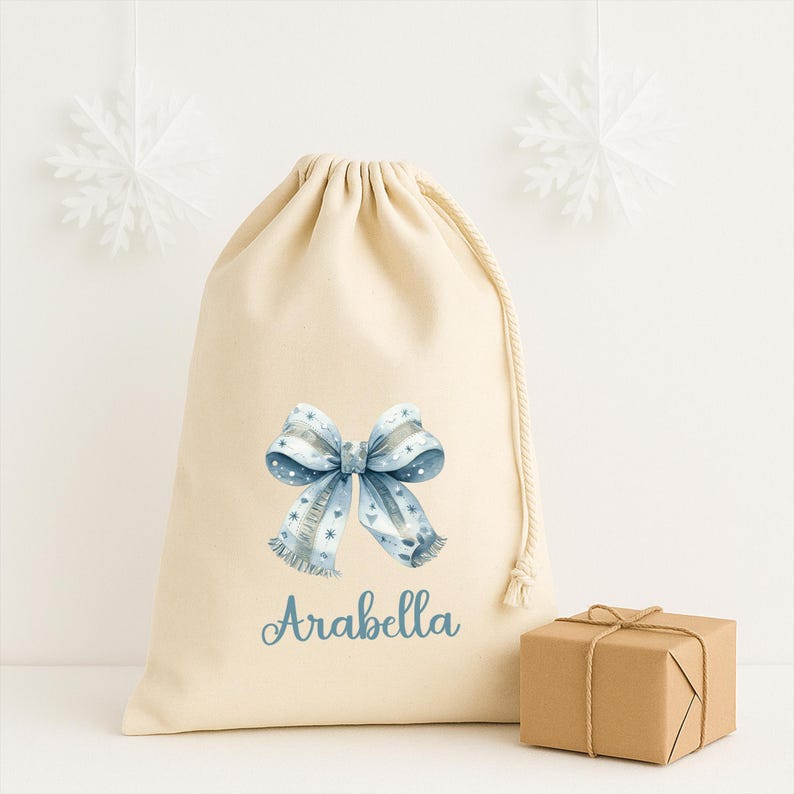 Puede incluir: Una bolsa de regalo beige con cord&oacute;n, con un dise&ntilde;o de lazo azul y el nombre "Arabella" impreso en azul. Una peque&ntilde;a caja de regalo envuelta est&aacute; a la derecha. Dos decoraciones de copos de nieve blancos cuelgan en el fondo.