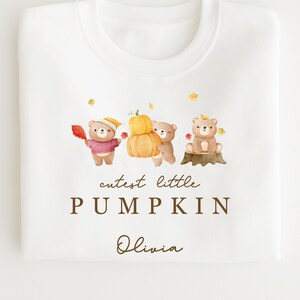 May include: White sweatshirt with a brown graphic of three bears holding a pumpkin and leaves. The text "cutest little PUMPKIN Olivia" is printed below the bears.