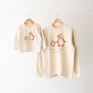 May include: Two cream colored sweatshirts with a graphic of two bears holding hands. The larger bear is wearing a pink bow. The text on the sweatshirts reads "Mama & Elsie since 2023".