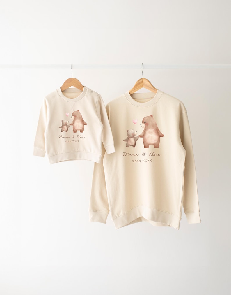 May include: Two cream colored sweatshirts with a graphic of two bears holding hands. The larger bear is wearing a pink bow. The text on the sweatshirts reads "Mama & Elsie since 2023".