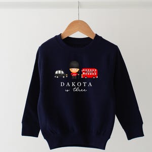 May include: Navy blue long-sleeved sweatshirt with a graphic of a cartoon London bus, a black taxi, and a cartoon figure wearing a red uniform. The text "DAKOTA is three" is printed below the graphic.
