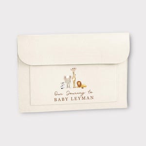 May include: Cream-coloured envelope-style baby announcement with a flap closure. The front pocket features watercolour illustrations of a zebra, elephant, giraffe, and lion. The text "Our Journey to BABY LEYMAN" is printed in brown.