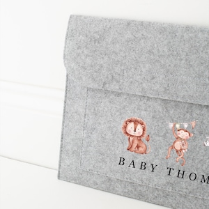 May include: A grey felt baby organiser with a flap closure. The organiser features watercolour illustrations of a lion and a monkey. The text "BABY THOM" is printed in black below the illustrations. The background is white.