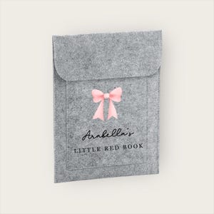 May include: A grey felt book cover featuring a pink bow and the text "Arabella's LITTLE RED BOOK". The cover has a flap closure and a front pocket. The design is simple and elegant.