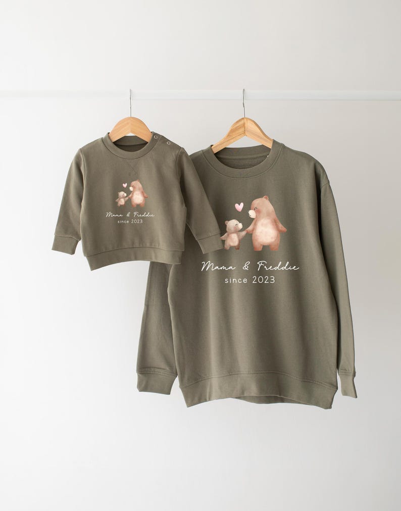 May include: Two green sweatshirts with a bear and cub design. The larger sweatshirt has the text "Mama & Freddie since 2023" printed on it. The smaller sweatshirt has the text "Mama & Freddie since 2023" printed on it.