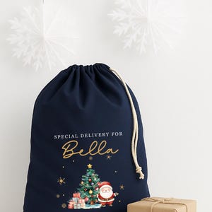 May include: A navy blue drawstring gift bag with the text "SPECIAL DELIVERY FOR Bella" in gold script. The bag features a Christmas tree, Santa Claus, and wrapped presents. A small wrapped gift sits beside the bag.