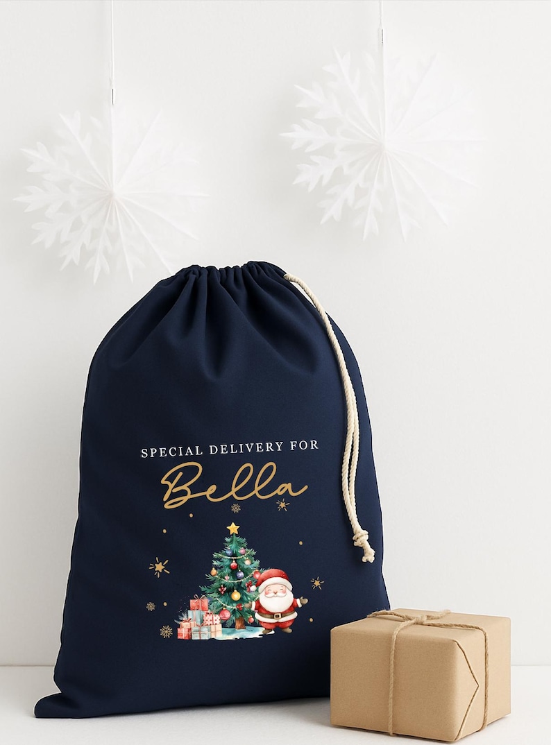 May include: A navy blue drawstring gift bag with the text "SPECIAL DELIVERY FOR Bella" in gold script. The bag features a Christmas tree, Santa Claus, and wrapped presents. A small wrapped gift sits beside the bag.