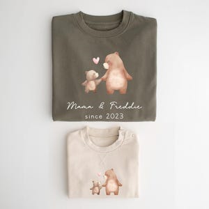 May include: Two brown bear illustrations on a green and beige sweatshirt set. The larger bear is holding the smaller bear's paw. The text "Mama & Freddie since 2023" is printed on the green sweatshirt.