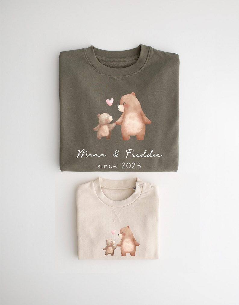 May include: Two brown bear illustrations on a green and beige sweatshirt set. The larger bear is holding the smaller bear's paw. The text "Mama & Freddie since 2023" is printed on the green sweatshirt.