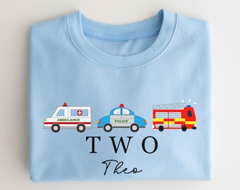 Puede incluir: Sudadera azul claro con ilustraciones de una ambulancia, un coche de policía y un camión de bomberos. El texto "TWO" y el nombre "Theo" están impresos debajo. Adecuado para un cumpleaños infantil.