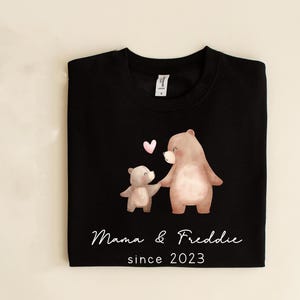 May include: Black sweatshirt with a design of two brown bears holding hands with a pink heart above them. The text "Mama & Freddie since 2023" is printed below the bears.