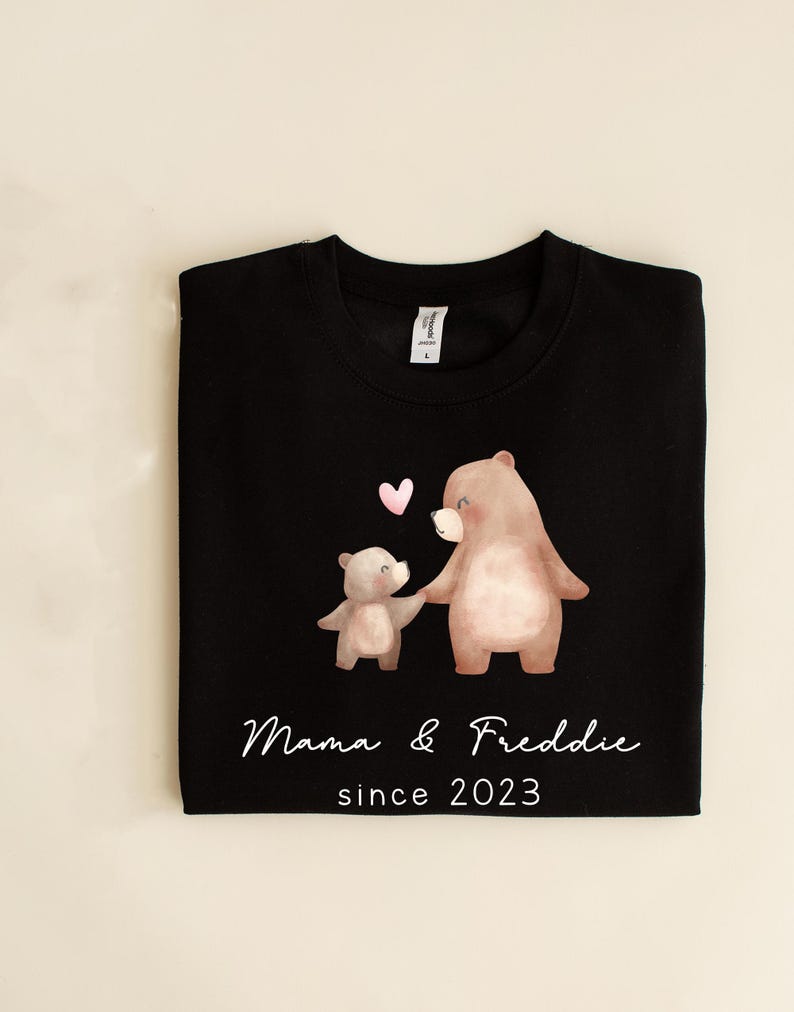 May include: Black sweatshirt with a design of two brown bears holding hands with a pink heart above them. The text "Mama & Freddie since 2023" is printed below the bears.