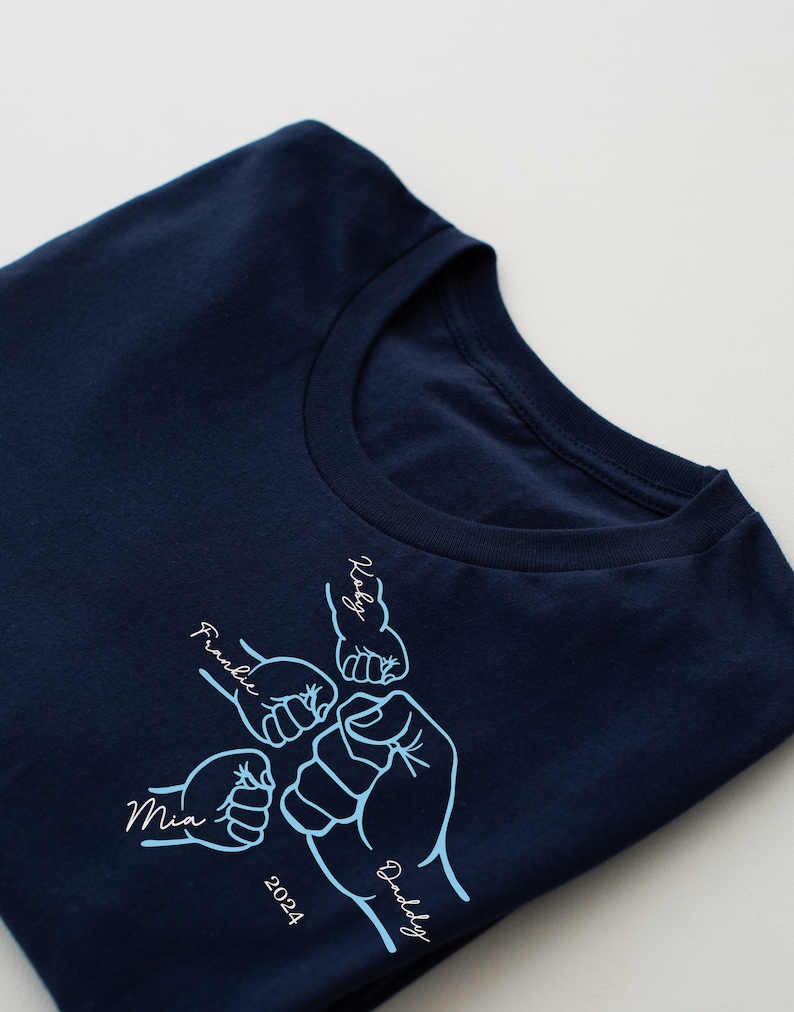 May include: A navy blue t-shirt with a design of five fists bumping each other. The fists are labeled with the names "Mia", "Frankie", "Daddy", "Koby", and the year "2024".