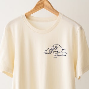 May include: A cream colored t-shirt with a graphic of two fists bumping, one labeled "Harper" and the other "Daddy" with the year "2024" underneath.