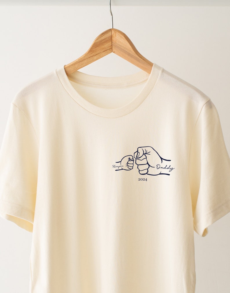 May include: A cream colored t-shirt with a graphic of two fists bumping, one labeled "Harper" and the other "Daddy" with the year "2024" underneath.