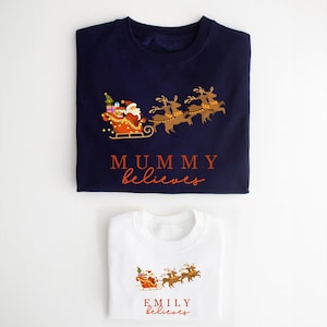 May include: Two folded Christmas sweaters, one navy blue and one white, with a Santa Claus and reindeer design. The navy blue sweater says "MUMMY believes" and the white sweater says "EMILY believes".