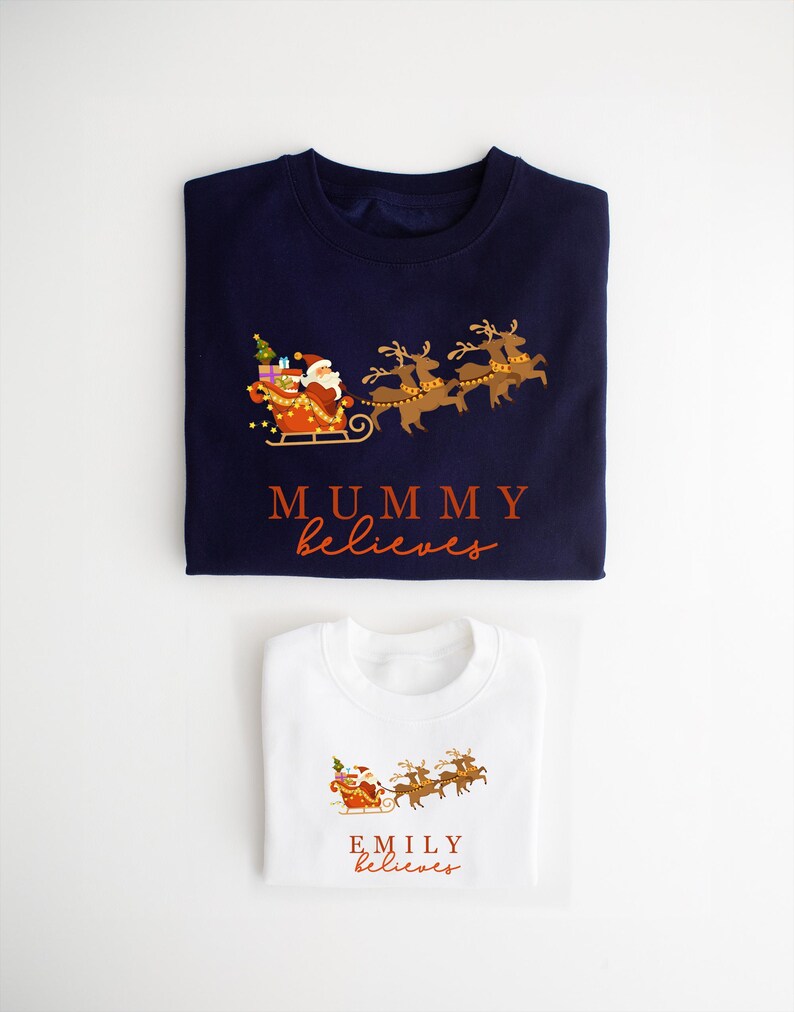 May include: Two folded Christmas sweaters, one navy blue and one white, with a Santa Claus and reindeer design. The navy blue sweater says "MUMMY believes" and the white sweater says "EMILY believes".