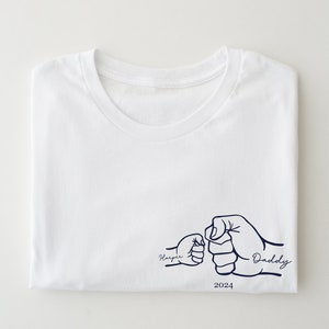 May include: White t-shirt with a blue graphic of two fists bumping, one labeled "Harper" and the other "Daddy" with the year 2024 below.