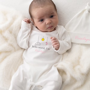 May include: A white babygrow with a graphic of a cloud, sun and moon with the text "hello world, I'm Penelope". The babygrow is worn by a baby lying on a white fluffy blanket. A white hat with the name "Penelope" embroidered in pink is lying on the blanket.
