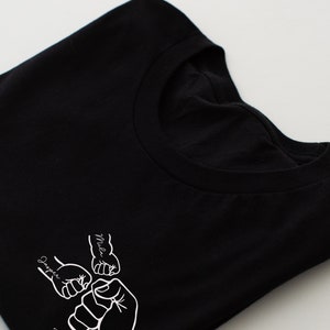 May include: Black t-shirt with a white graphic of four fists bumping together. The fists are labeled "Daddy", "Mila", "Harper", and "2024".