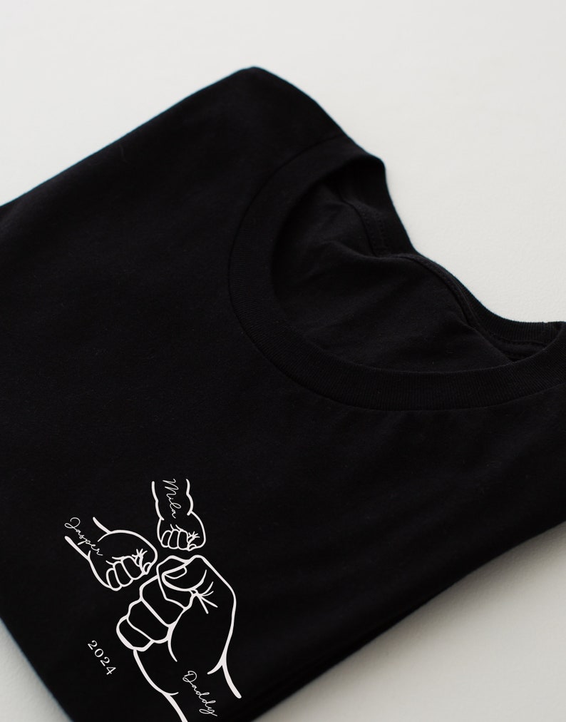 May include: Black t-shirt with a white graphic of four fists bumping together. The fists are labeled "Daddy", "Mila", "Harper", and "2024".