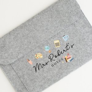 May include: Grey felt laptop sleeve with a pocket and a design featuring a coffee cup, calculator, globe, paint palette, pencils, books, and stars. The text "Mrs. Roberts' notes" is written in black cursive.