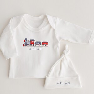 May include: White long-sleeved baby onesie with a red and blue train graphic and the word "ATLAS" printed below. A matching white baby hat with the word "ATLAS" printed on the front.