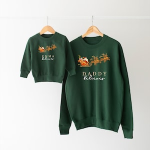 May include: Two green Christmas sweaters with Santa Claus and reindeer graphics. The smaller sweater says "Emma believes" and the larger sweater says "Daddy believes".
