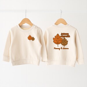 May include: Cream-coloured sweatshirt with long sleeves, featuring a cartoon leaf design on the front and back. The back of the sweatshirt has the text "Autumn days with Mummy & Aurora" above the leaf design. The sweatshirt is displayed on a wooden hanger.