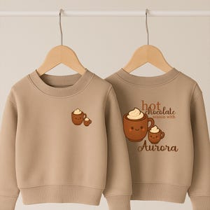 May include: Two beige sweatshirts featuring a hot chocolate design. One has a small graphic of two hot chocolate mugs with cream. The other has a larger graphic with the text "hot chocolate season with Aurora."