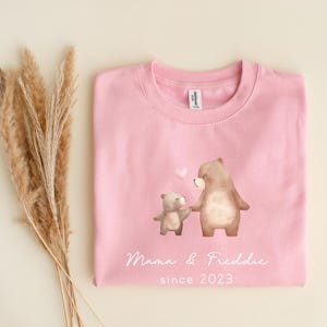 May include: Pink sweatshirt with a graphic of two brown bears holding hands. The text "Mama & Freddie since 2023" is printed below the bears.