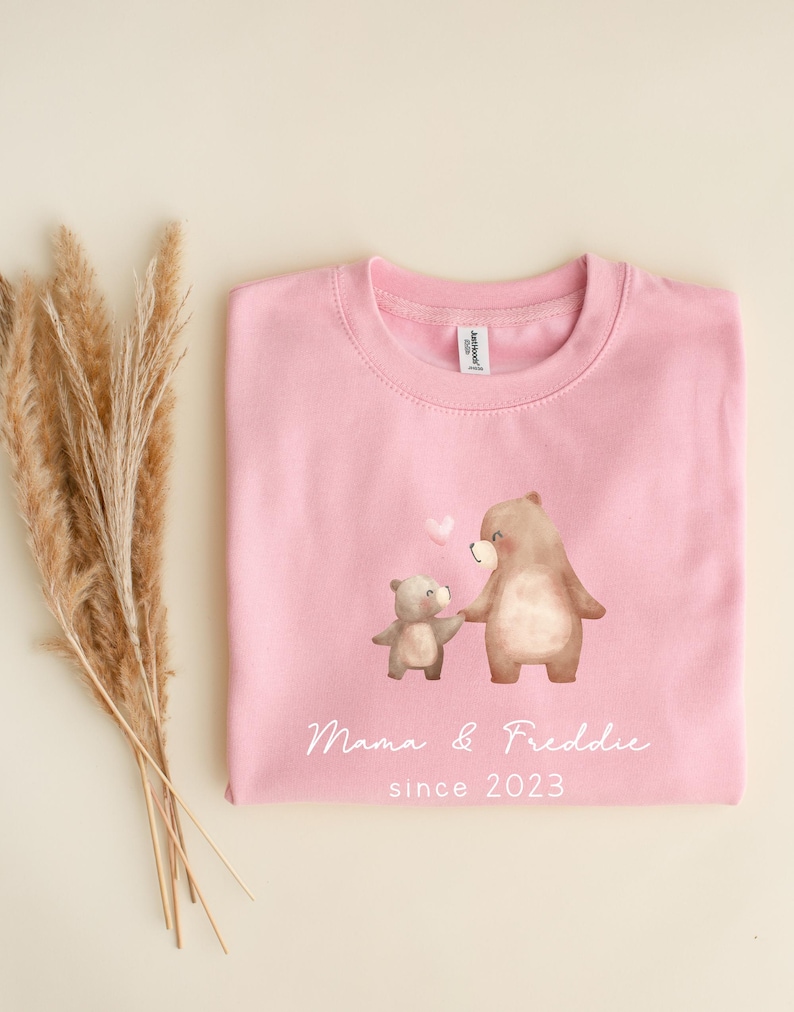 May include: Pink sweatshirt with a graphic of two brown bears holding hands. The text "Mama & Freddie since 2023" is printed below the bears.