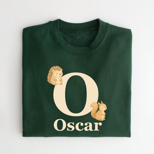 May include: Dark green sweatshirt with the letter "O" and the name "Oscar" in white. A hedgehog and a squirrel are on either side of the letter. The sweatshirt is folded.