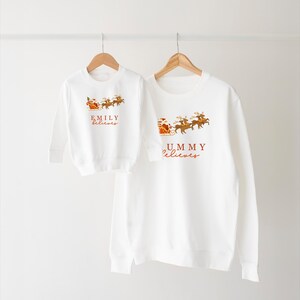 May include: White Christmas themed long-sleeve sweatshirts with Santa Claus and reindeer graphics. The larger sweatshirt says "Mommy Believes" and the smaller sweatshirt says "Emily Believes".