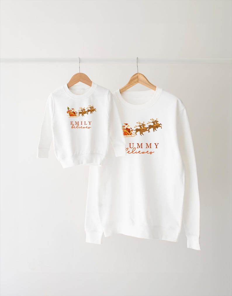 May include: White Christmas themed long-sleeve sweatshirts with Santa Claus and reindeer graphics. The larger sweatshirt says "Mommy Believes" and the smaller sweatshirt says "Emily Believes".