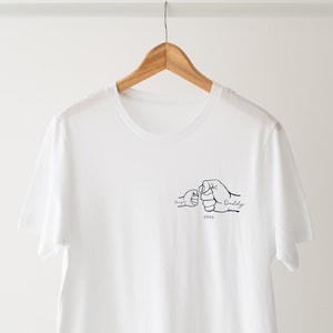 May include: White t-shirt with a navy blue design of two fists bumping, one labeled "Daddy" and the other "My Guy", with the year "2024" below.
