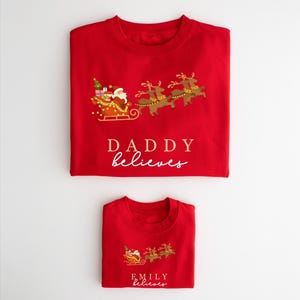 May include: Two red Christmas shirts with a Santa Claus and reindeer design. The larger shirt says "Daddy believes" and the smaller shirt says "Emily believes".