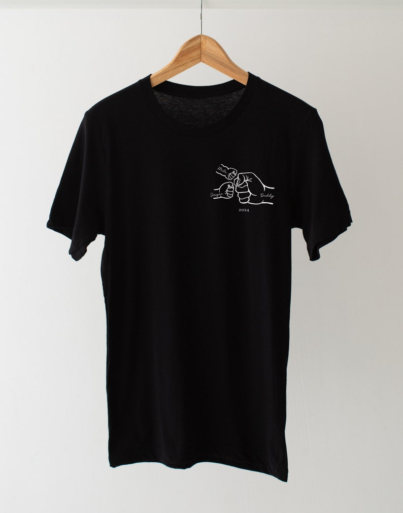 May include: Black t-shirt with a white graphic of four hands clasped together, labeled "Mommy", "Daddy", "Sister", and "Buddy". The year "2023" is printed below the hands.