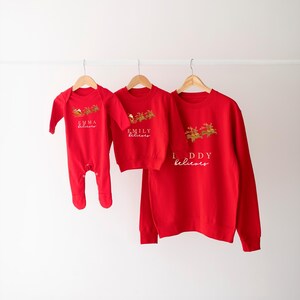 May include: Red Christmas sweaters with the text "Emma believes", "Emily believes", and "Daddy believes" on them. The sweaters are hanging on hangers against a white wall.