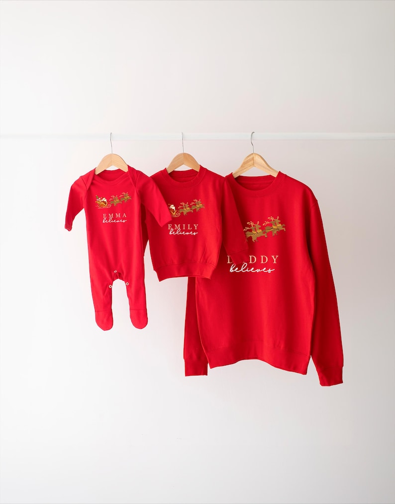 May include: Red Christmas sweaters with the text "Emma believes", "Emily believes", and "Daddy believes" on them. The sweaters are hanging on hangers against a white wall.