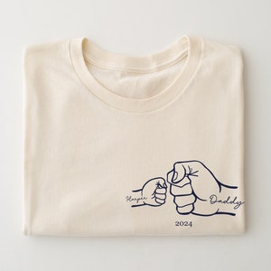 May include: A cream colored t-shirt with a graphic of two fists bumping, one labeled "Harper" and the other "Daddy". The year 2024 is printed below the graphic.